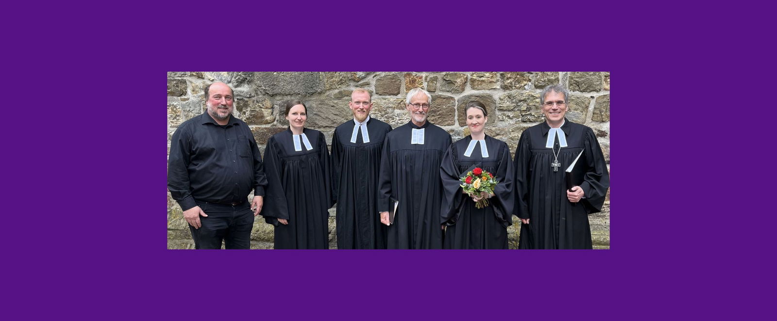 Sechs Personen stehen nebeneinander vor einer steinernen Wand. Fünf tragen Talar, einer ein dunkles Hemd. Eine weiblich gelesene Person hält einen Blumenstrauß in Händen.
