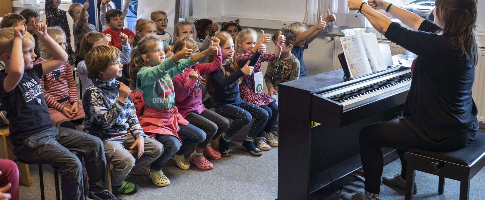 Eine als Frau lesbare Person gibt einer Schulklasse von einem Klavier aus ein Zeichen und hält beide Daumen hoch.