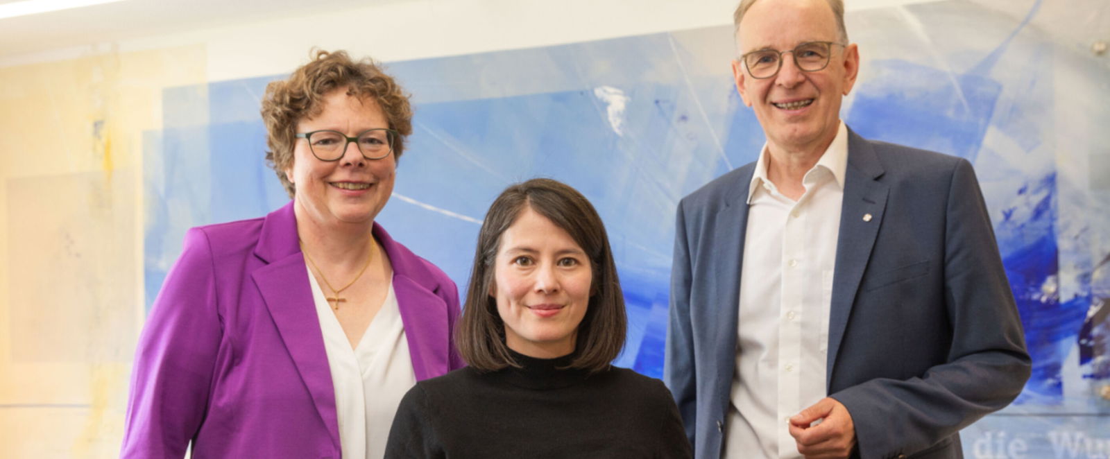 drei Menschen vor einem blau-gelben Aquarellbild schauen in die Kamera und lächeln