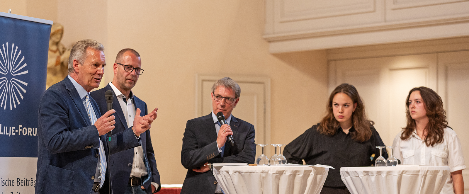 Drei männlich gelesene Personen und zwei weibliche stehen zusammen an weißen Tischen. Einer hält ein Mikro in der Hand.