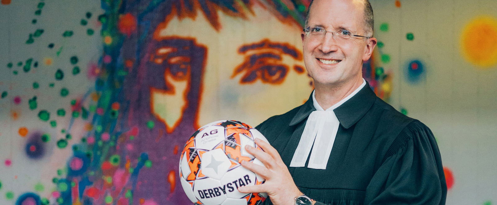 Eine als Mann lesbare Person im Talar mit einem Fußball vor einem Jesus-Graffito