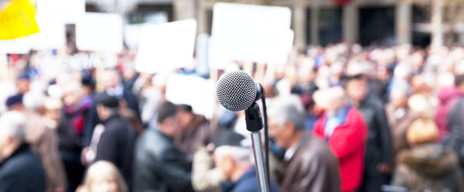 Ein Mikrofon. Im Hintergrund demonstrierende Menschen.