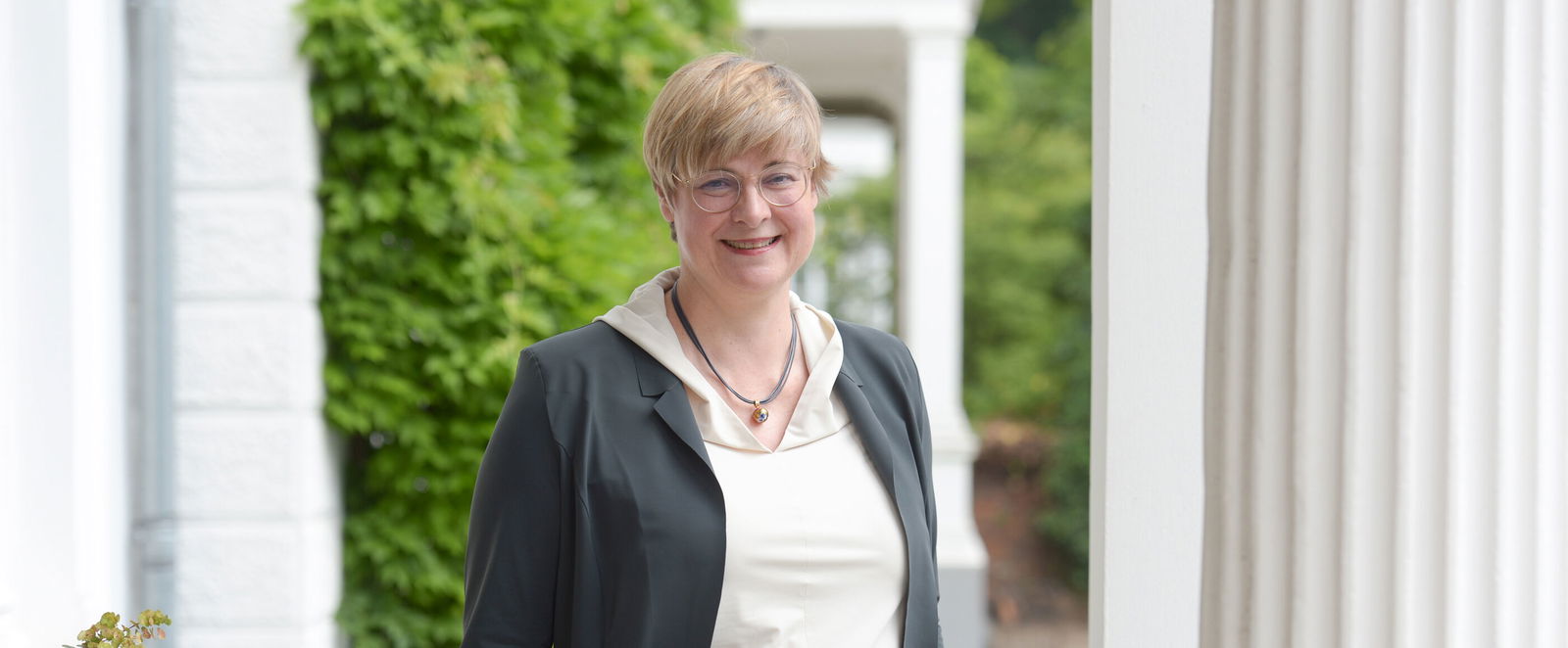 Eine als Frau lesbare Person steht im Freien zwischen weißen Säulen.