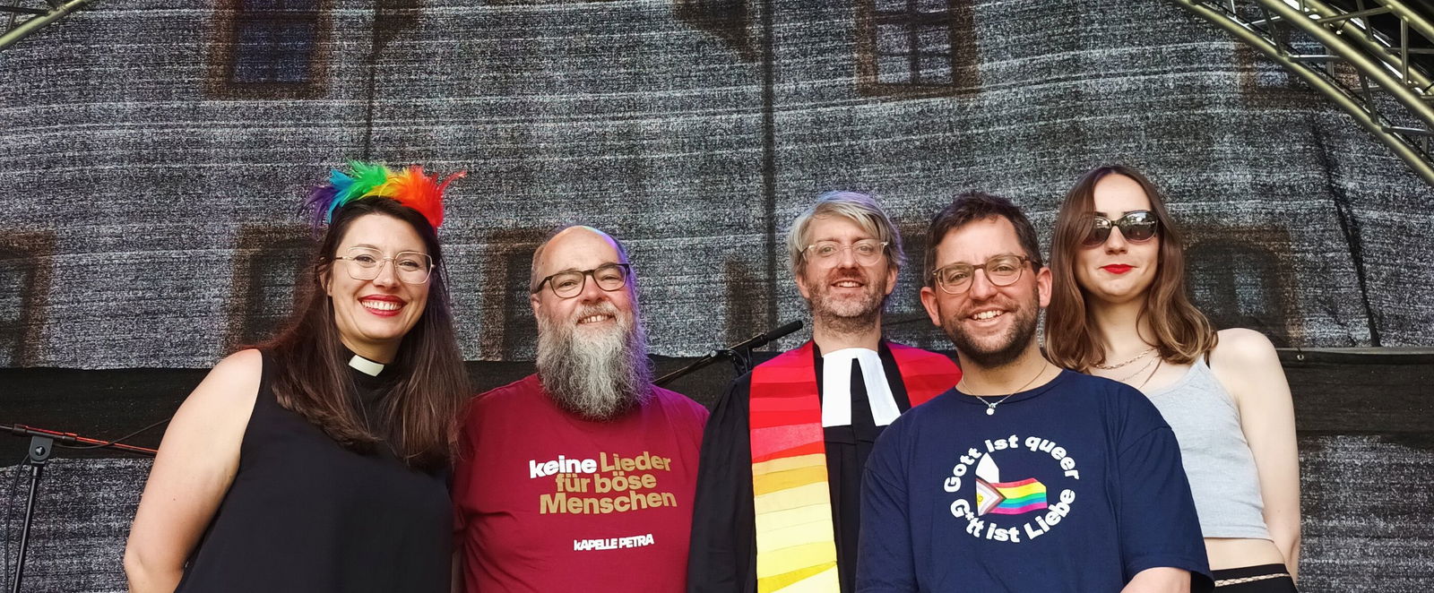 Fünf Menschen auf einer Bühne mit bunten Scheinwerfern im Hintergrund. Manche tragen regenbogen-bunte Accessoires.