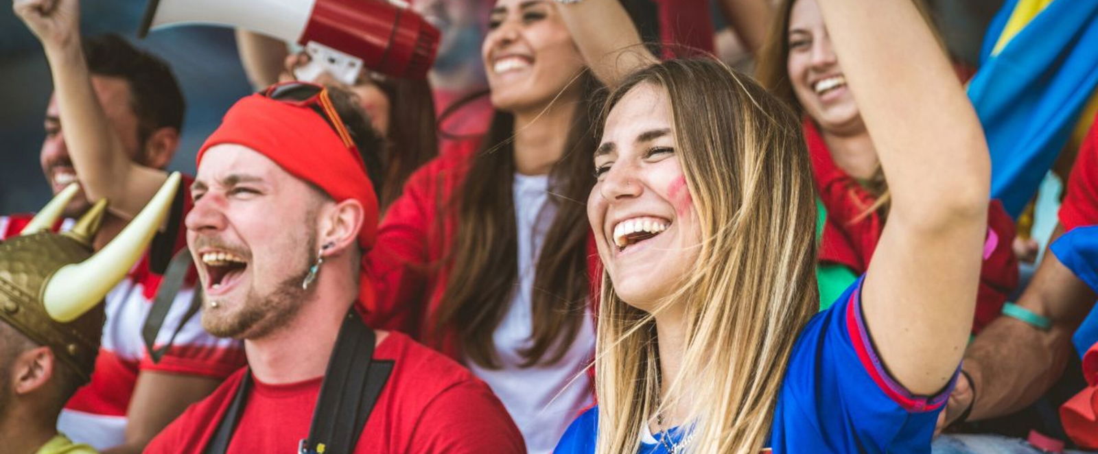 Weiblich und männliche gelesene Personen mit bunten Trikots und Accessoires stehen auf einer Tribüne und jubeln.