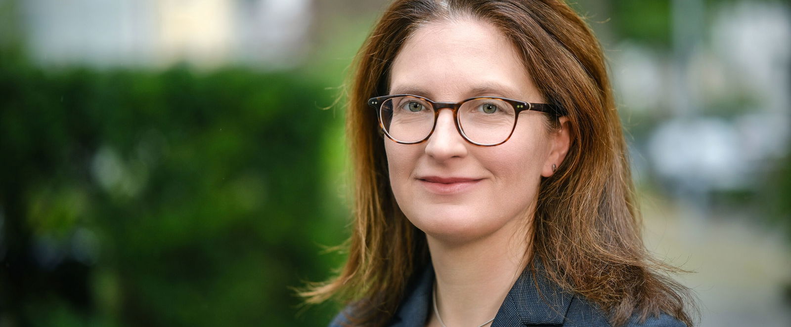 Eine weiblich gelesene Person mit Brille und mittellangen, braunen Haaren und Brille in blauem Jackett steht draußen vor einer grünen Hecke.