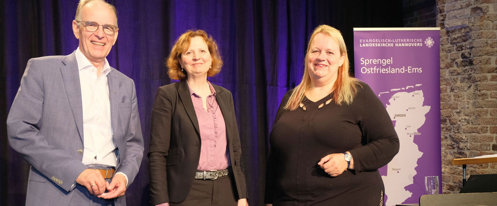 Eine als Mann und zwei als Frauen lesbare Personen posieren vor einem Aufsteller des Spremgels Ostfriesland-Ems