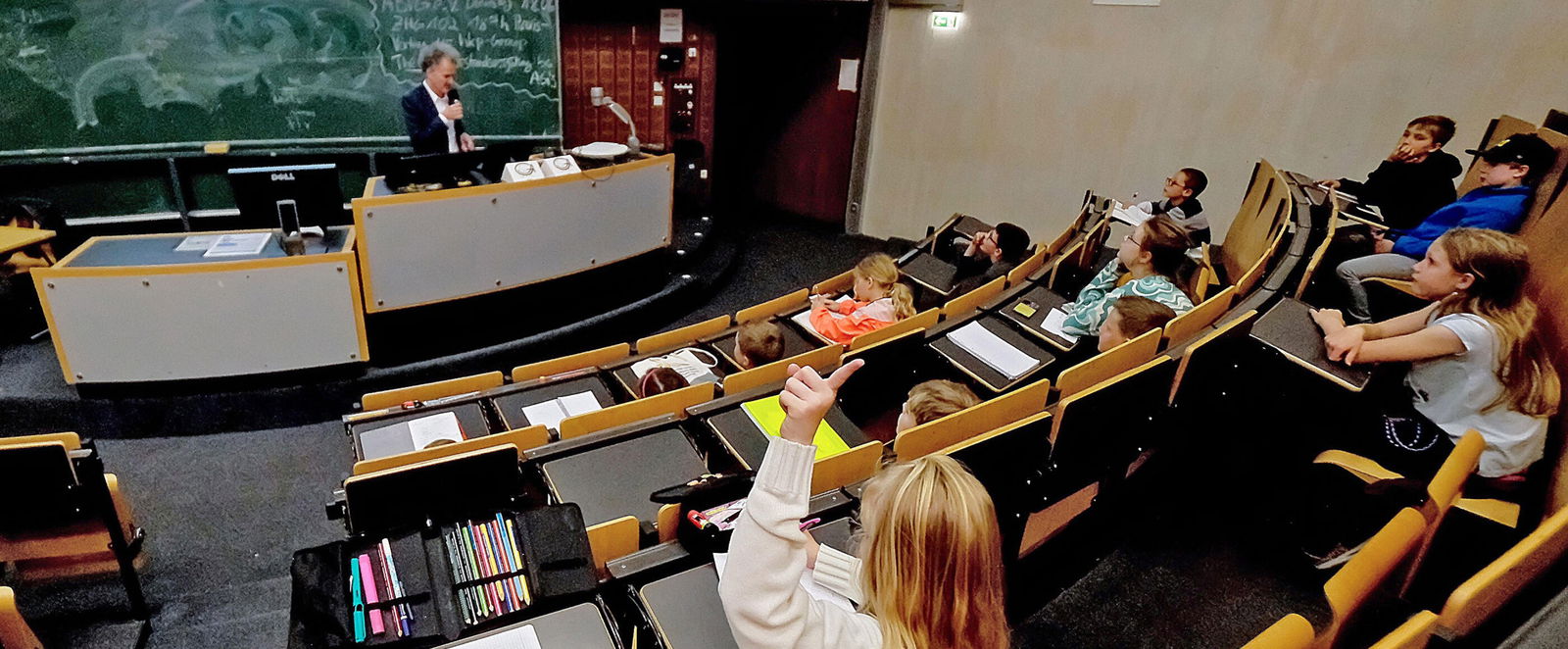 Kinder hören in einem Hörsaal einem Professor zu. Eine als Mädchen lesbare Person meldet sich.