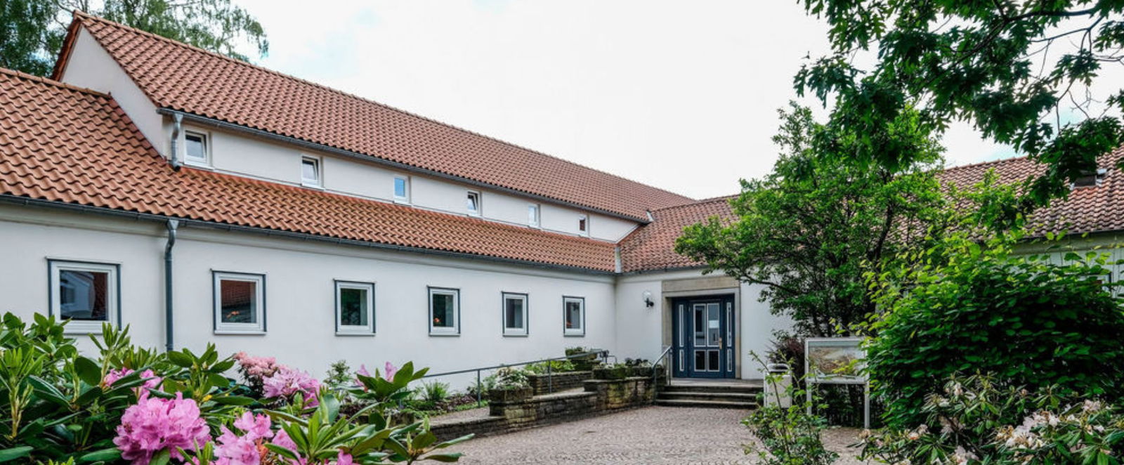 Ein weißes Gebäude mit Ziegeldach, davor stehen Bäume und ein Busch mit rosafarbenen Blumen.