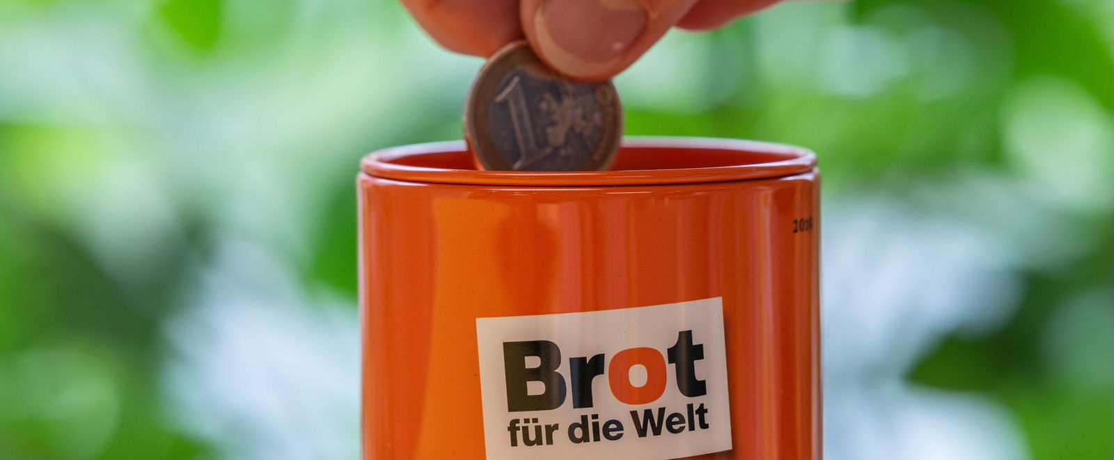 Orangfarbene Spendendose mit dem Logo von Brot für die Welt, eine Hand steckt eine Euromünze hinein.