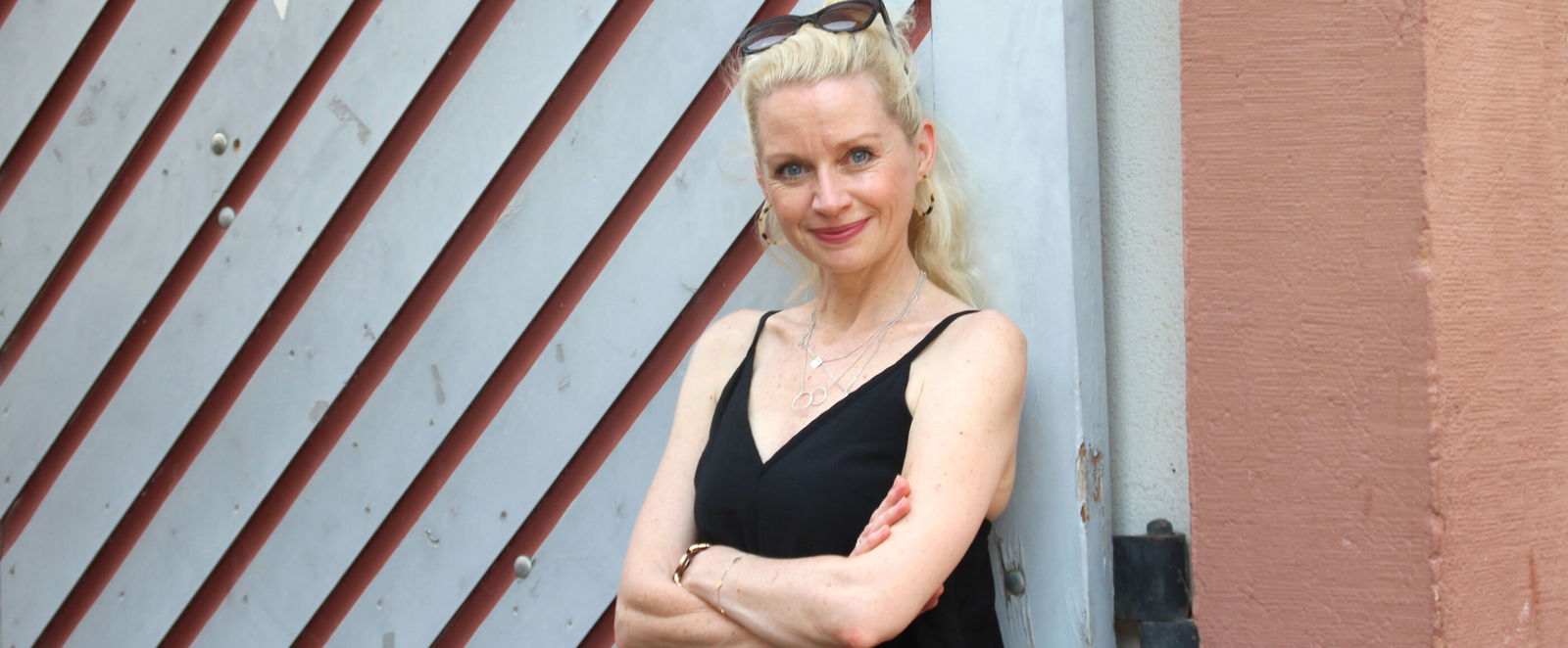 Eine blonde Frau trägt ein schwarzes Kleid und hat die Arme verschränkt.