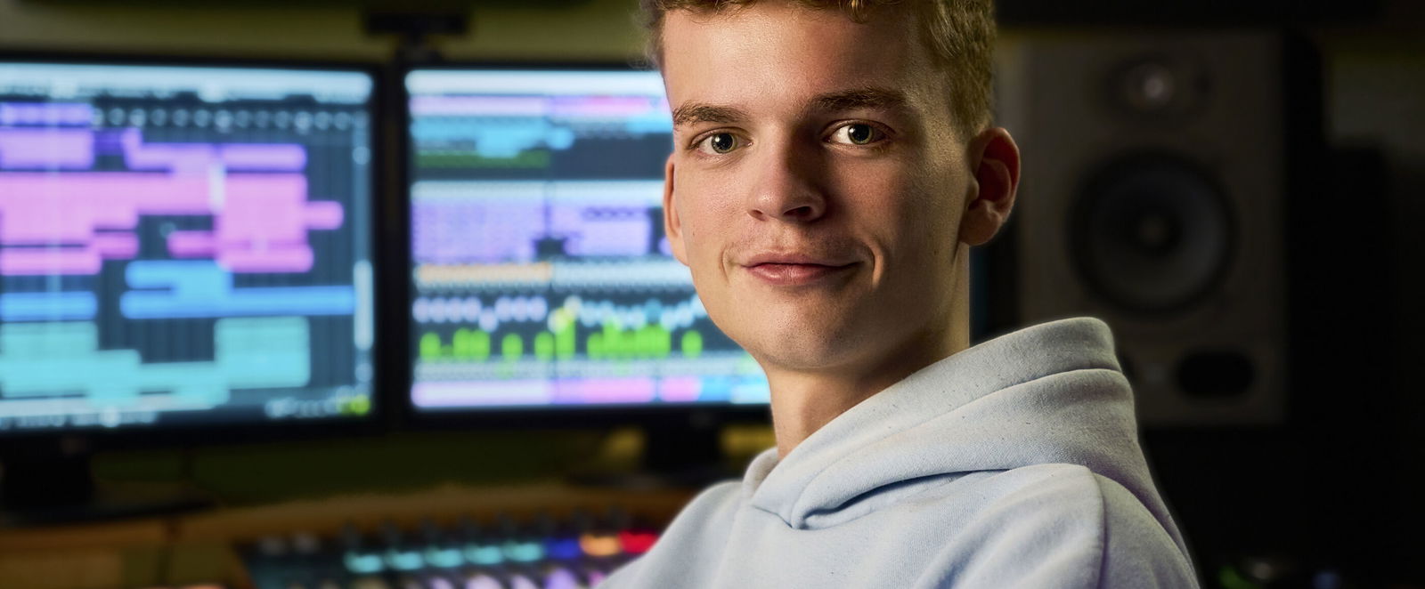 Eine männlich gelesene Person mit hellblauem Hoodie sitzt an einem Mischpult und Bildschirmen.