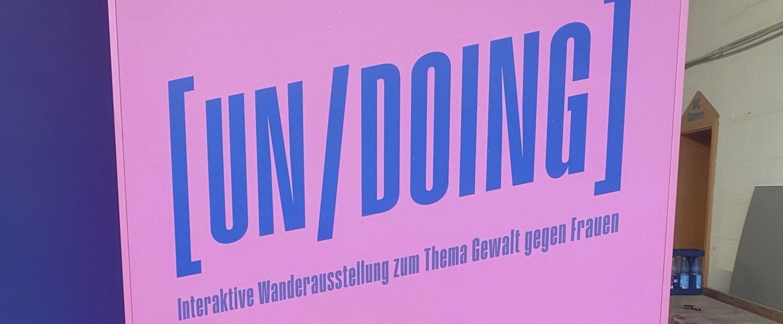 Blau-lila steht auf einem rosa Plakat: (Un / Doing) Interaktive Wanderausstellung über Gewalt gegen Frauen