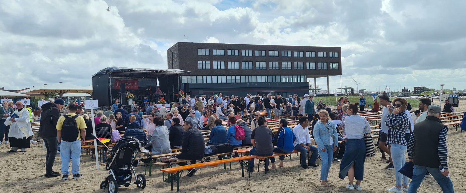Auf einem Strand sitzen Menschen auf Bierbänken vor einer Bühne, im Hintergrund ein großes Haus. Manche Menschen stehen oder laufen umher.