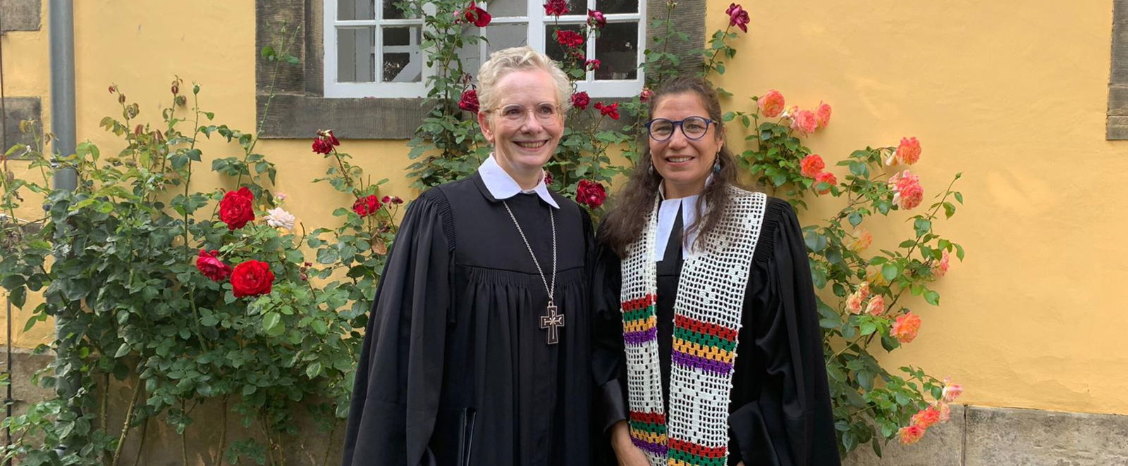 Zwei weiblich gelesene Personen, beide im Talar, stehen lächelnd vor einer gelben Hauswand mit roten Blumen.