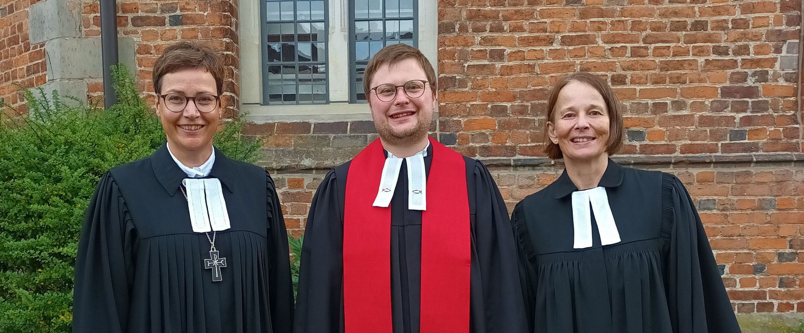 Zwei weiblich gelesene Personen und eine männlich gelesene stehen nebeneinander vor einer Backsteinkirche, alle tragen Talare. Die mittlere Person auch einen roten Schal.