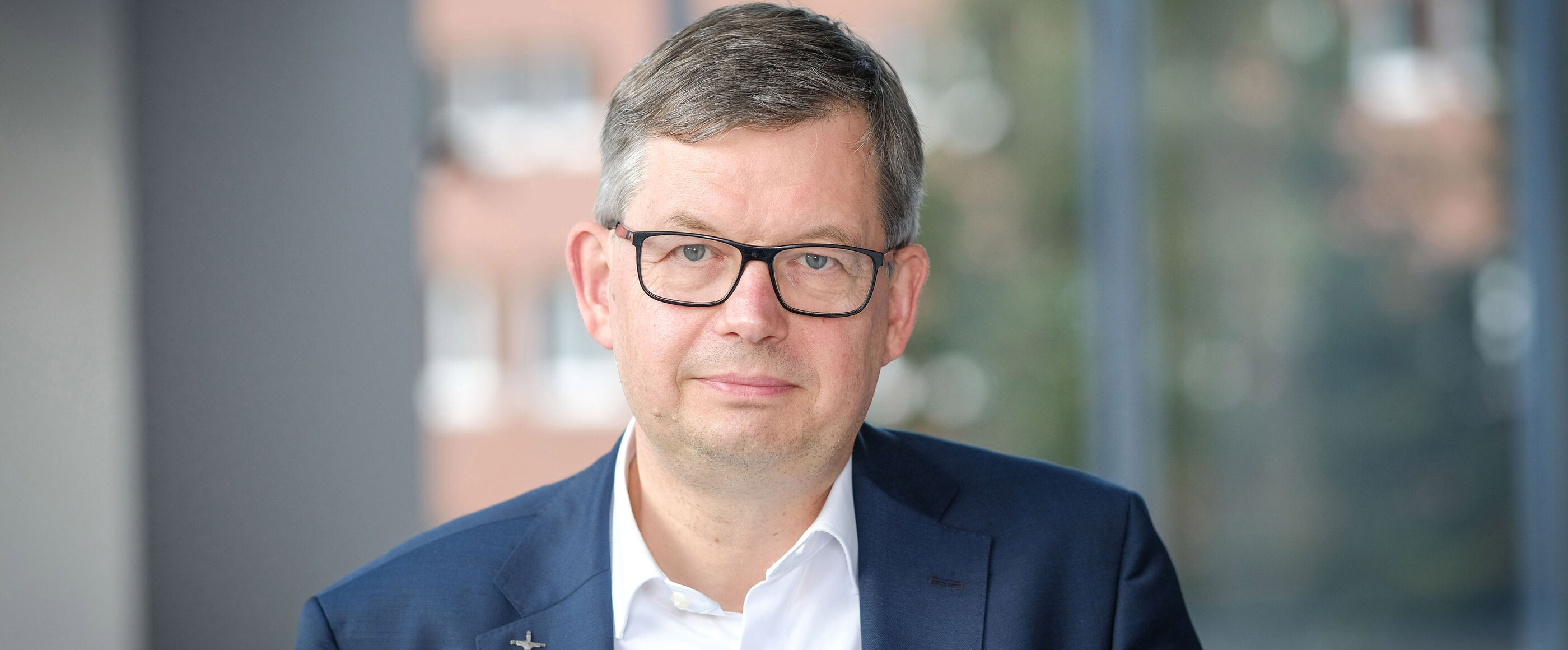 Eine als Mann lesbare Person mit blauem Sakko, weißem Hemd und Brille.