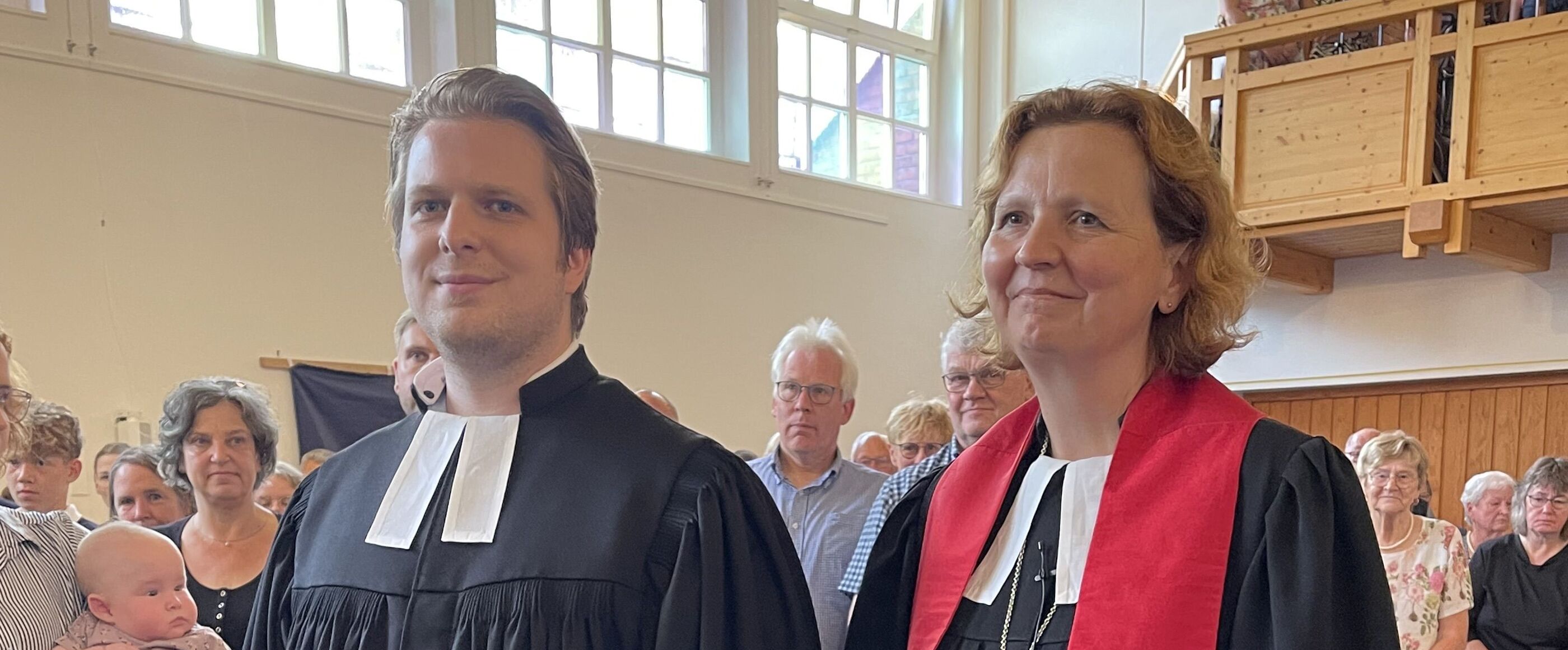 Eine männlich und eine weiblich gelesene Person, beiden in schwarzen Talare, gehen durch den Mittelgang einer Kirche zwischen stehenden Menschen hindurch.