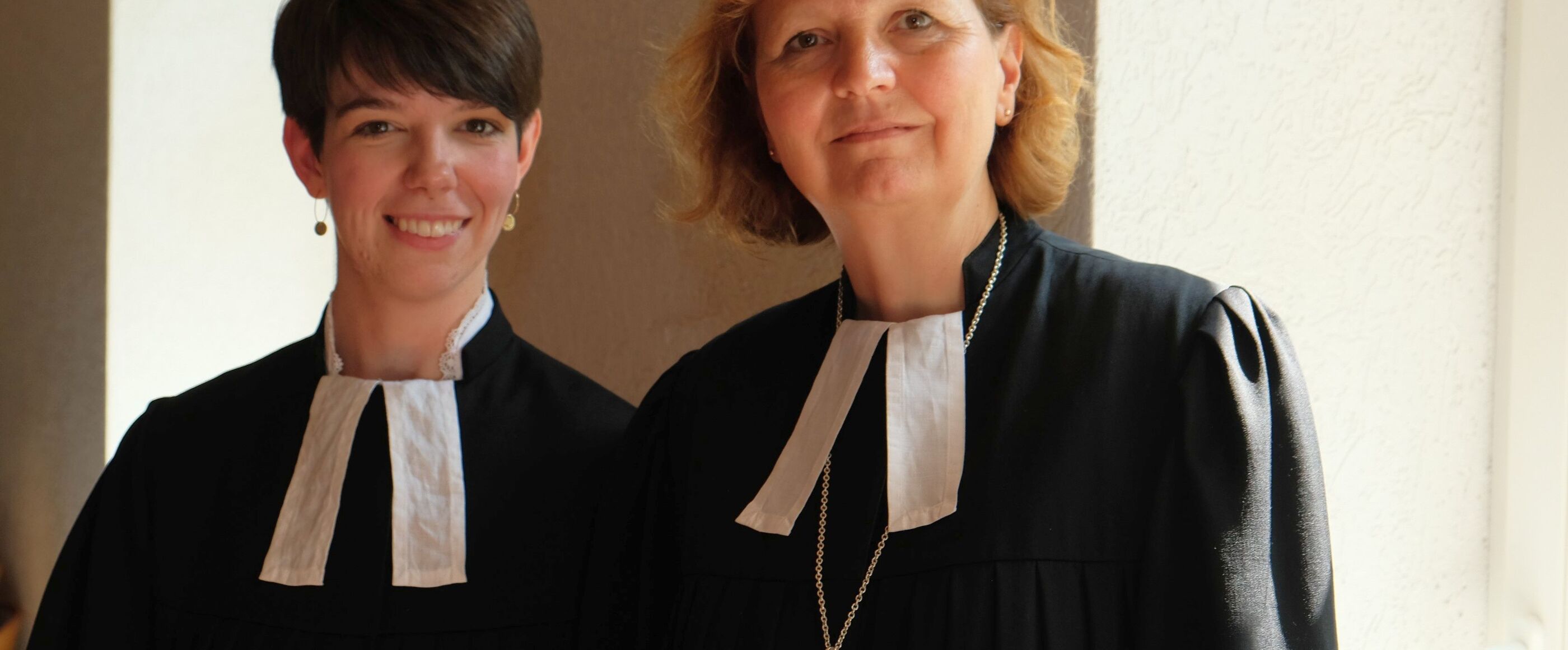 Zwei weiblich gelesene Personen mit Talaren stehen nebeneinander und lächeln.