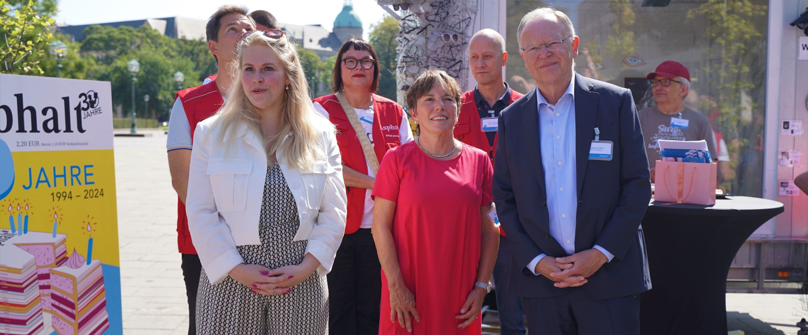 eine Gruppe von Menschen vor einem Gebäude. Links ein Plakat. Darauf ist ein Magazincover.