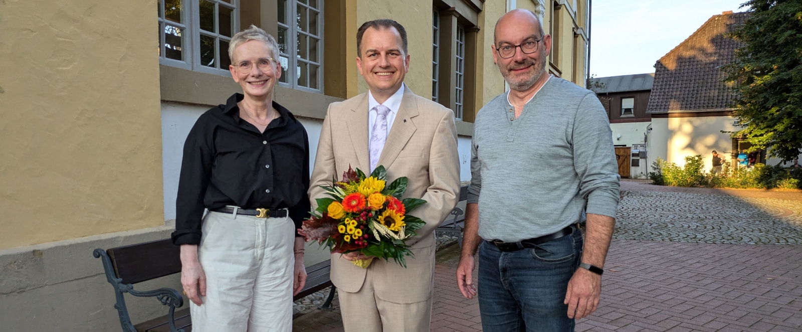 Eine als Frau und eine als Mann lesbare Person rahmen eine als Mann lesbare Person mit Blumenstrauß.
