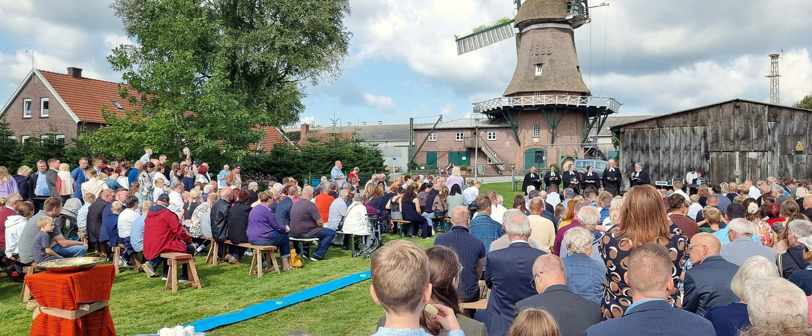 Auf einer Wiese sitzen viele Menschen auf Bänken und blicken nach vorn. Im Hintergrund eine Windmühle vor blauem Himmel mit weißen Wolken.