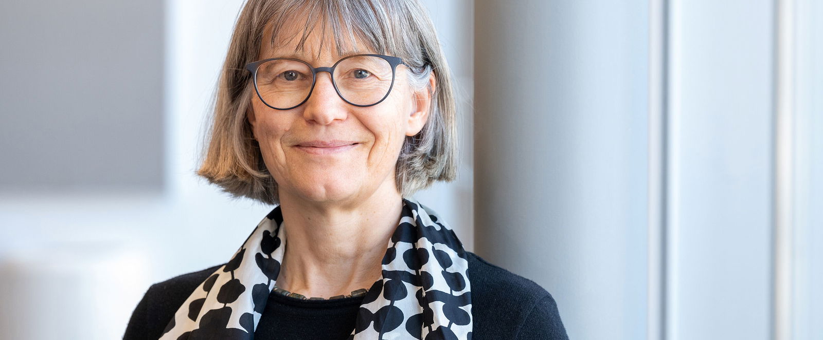 Eine als Frau lesbare Person mit halblangen grauen Haaren, Brille und gepunktetem Halstuch.