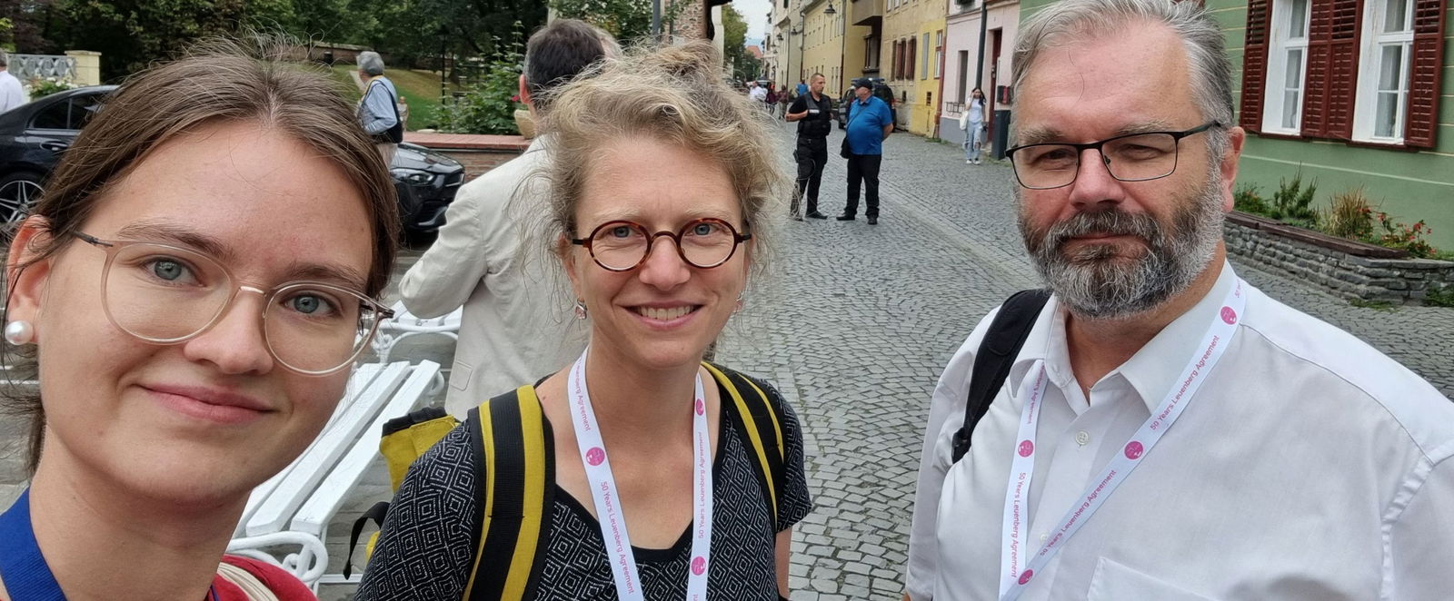 Eine männlich gelesene und zwei weiblich gelesene Personen stehen in einer Stadt.