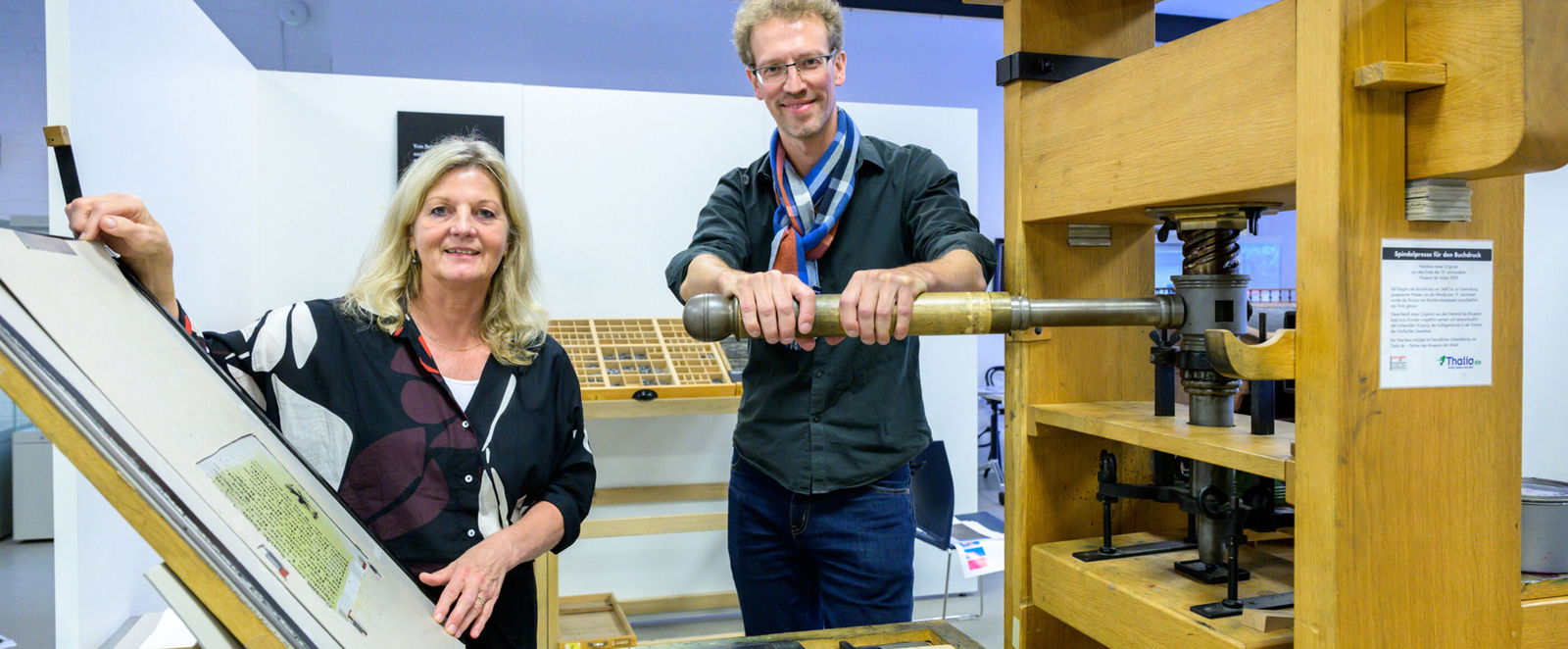 Eine als Frau und eine als Mann lesbare Person zeigen in einem Museum eine historische Druckmaschine.