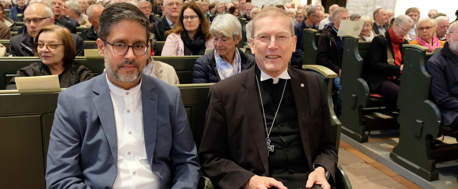 Zwei männlich gelesene Personen sitzen nebeneinander in einer Kirchenbank, viele andere Menschen ringsum. 