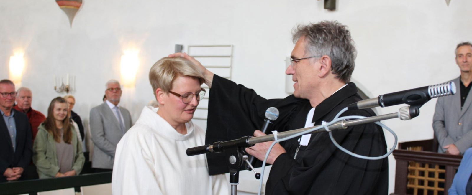 Eine weiblich gelesene Person in weißem Talar bekommt eine Hand von einer männlich gelesenen Person in schwarzem Talar auf den Kopf gelegt. 