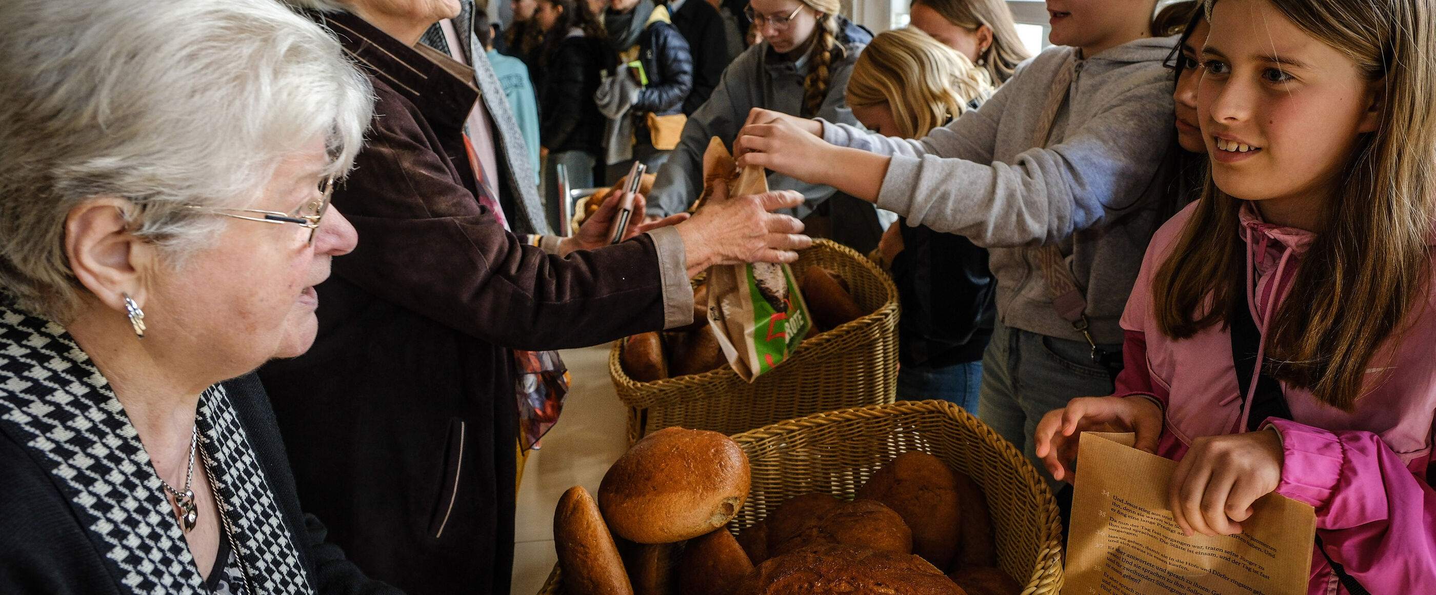 Jüngere Menschen stehen rechte von Brotkörben und bieten sie offenbar zum Kauf an. Links stehen ältere Menschen, eine Person nimmt gerade ein Brot entgegen.