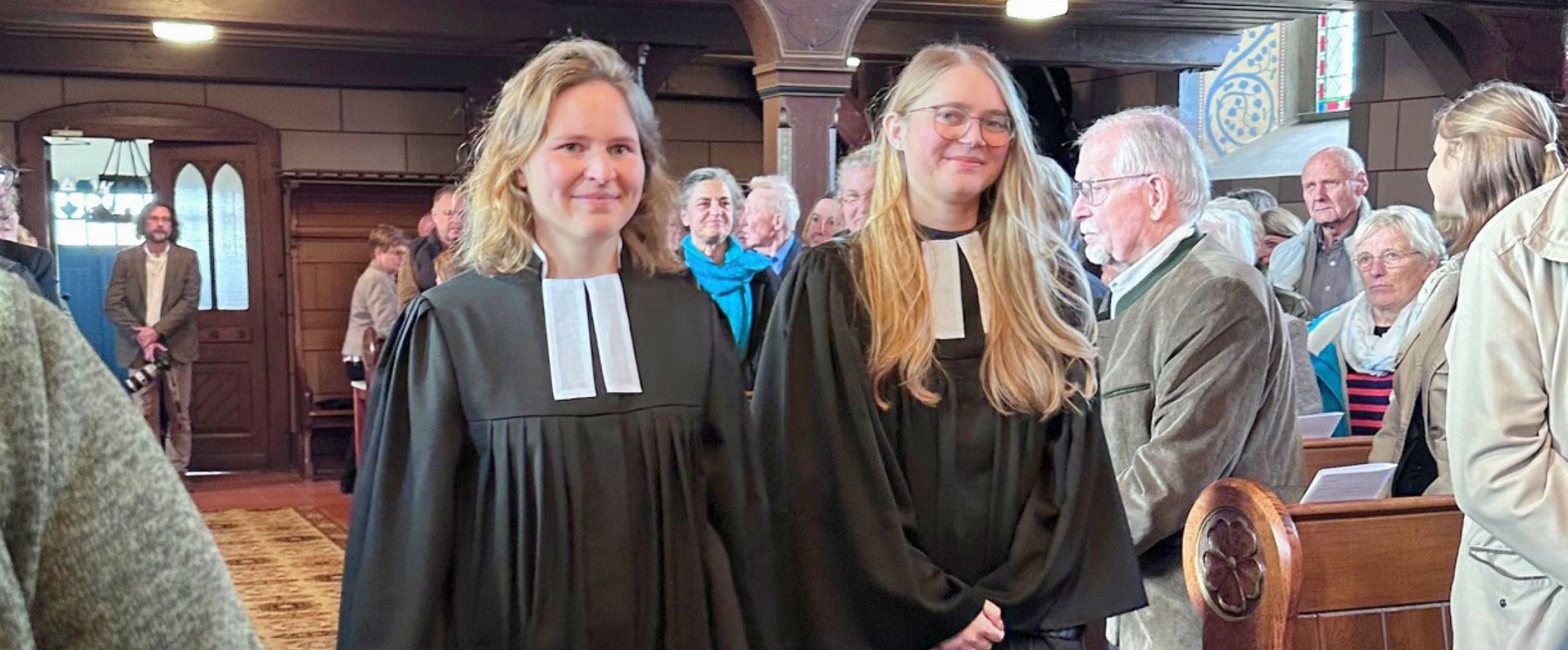 Zwei weiblich gelesene Personen im Talar stehen in einer geschmückten Kirche. Sie lächeln und haben beide eine Bibel in der Hand.