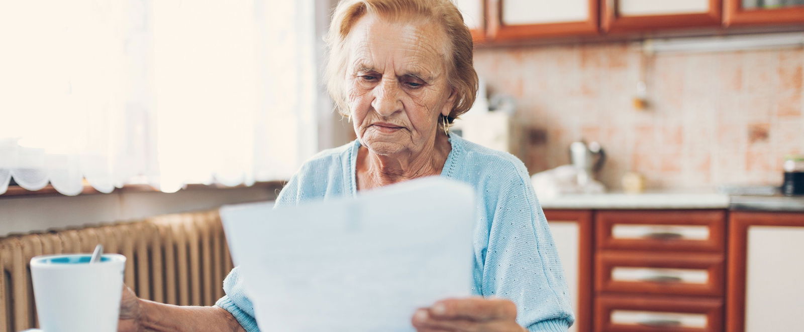Eine Seniorin sitzt in der Kueche und schaut besorgt auf einen Brief in ihrer Hand.
