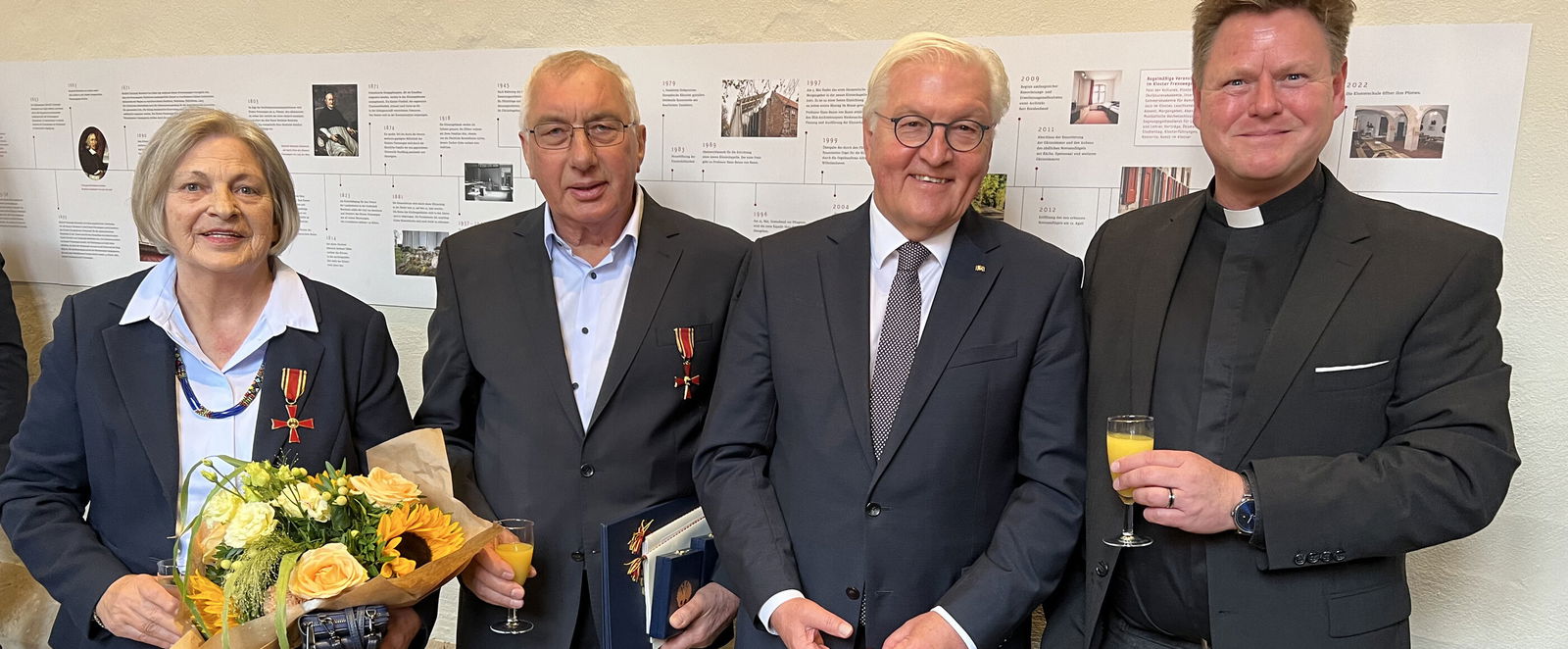 Eine weiblich gelesene Person hält einen Blumenstrauß und trägt das Bundesverdienstkreuz. Daneben drei männlich gelesene Personen, eine von ihnen trägt auch das Bundesverdienstkreuz. Die dritte Person von rechts ist der Bundespräsident.