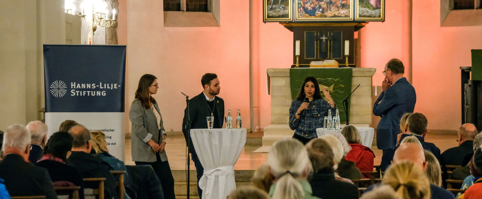 Vier Personen stehen in einem Halbkreis vor in Bänken sitzenden Menschen in einer Kirche.