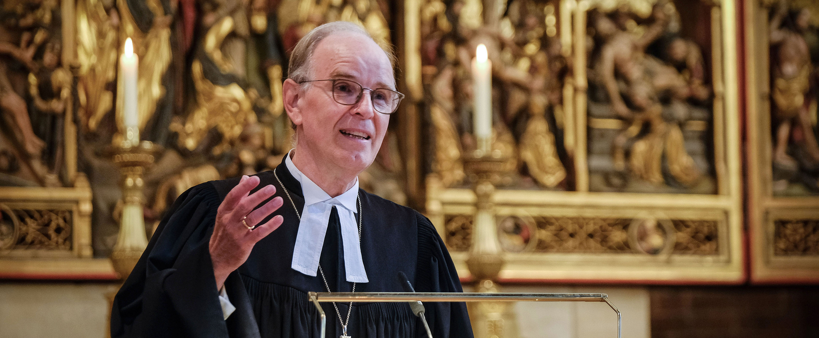 Eine männlich gelesene Person mit wenig Haar, Brille, im Talar spricht an einem Redepult vor einem goldenen Altar.