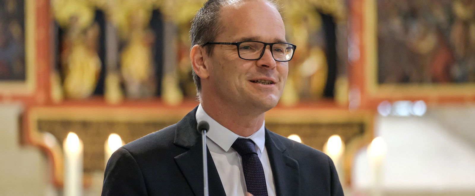 Eine als Mann lesbare Person im Anzug und mit hoher Stirn spricht in einer Kirche an einem Pult.