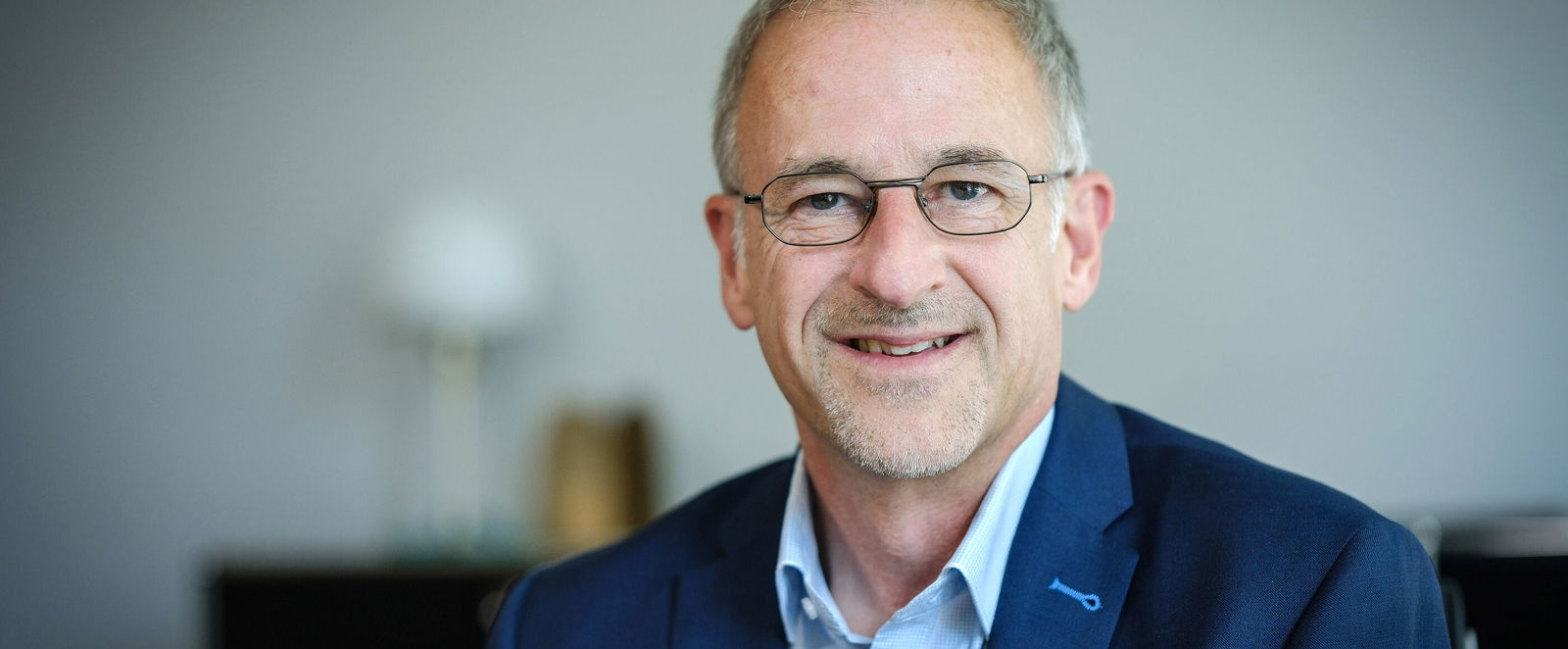 Eine als männlich lesbare Person im hellblauen Hemd, mit dunkelblauem Hemd und Brille vor grünem Hintergrund.