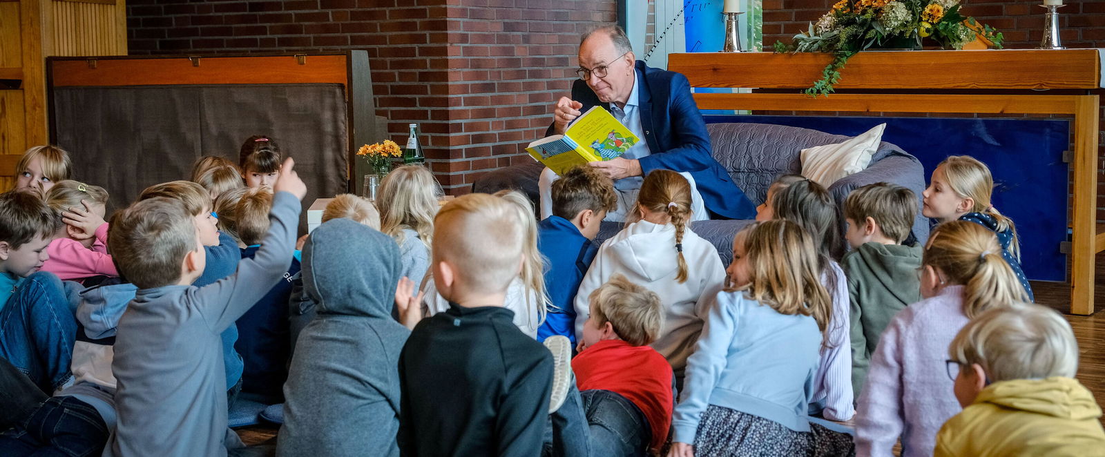 Eine männlich gelesene Person sitzt vor einer Gruppe Kinder und liest etwas aus einem Buch.