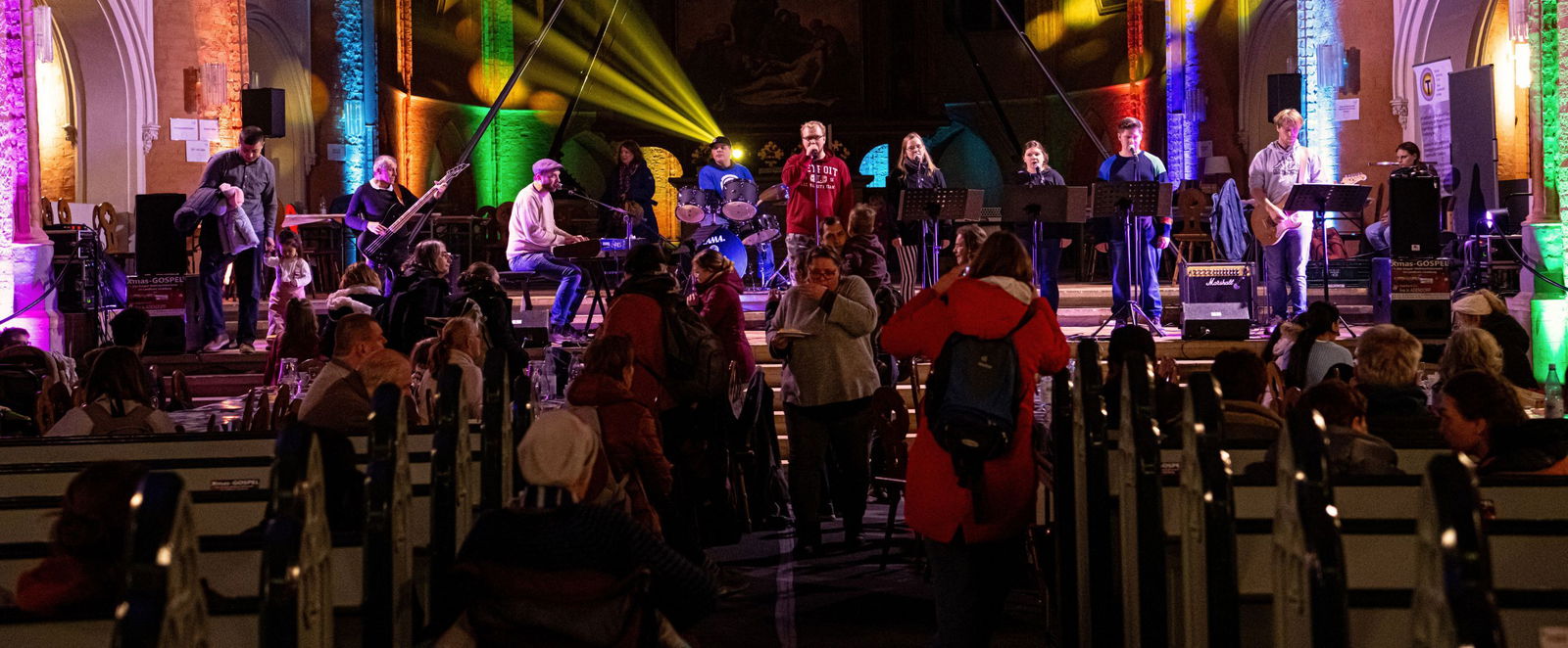 eine Band spielt in einer Kirche vor Publikum. Bunte Scheinwerfer strahlen in den Kirchraum.