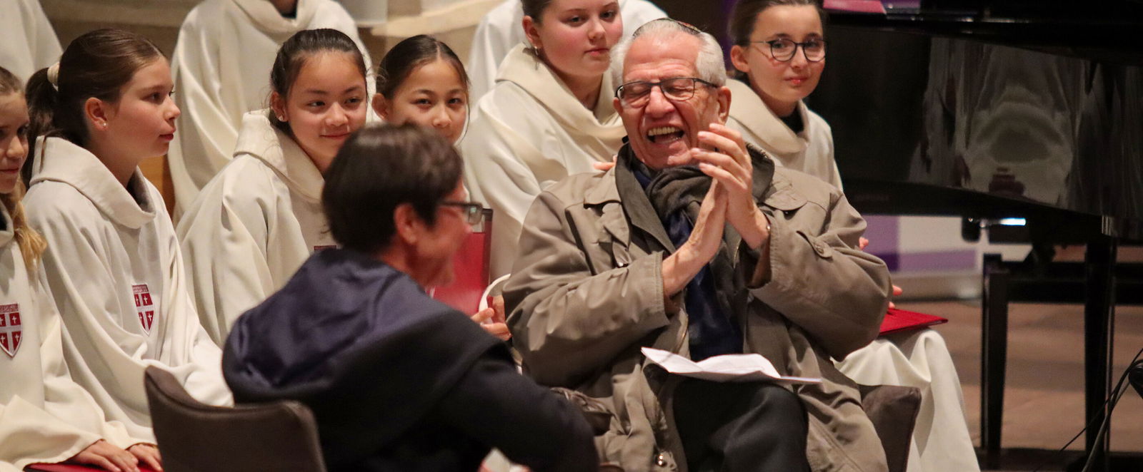 Zwei Personen, eine weiblich und eine männlich gelesene, sitzen nebeneinander, sich leicht zugewandt. Im Hintergrund junge weibliche Personen in weißen Gewändern.