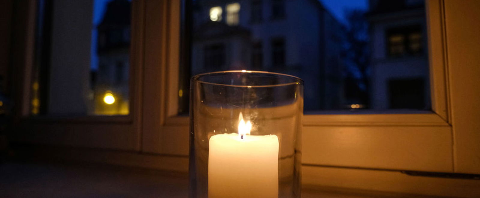 Eine Kerze brennt in einem Glas, das auf einer Fensterbank steht. Draußen ist es dunkel. 