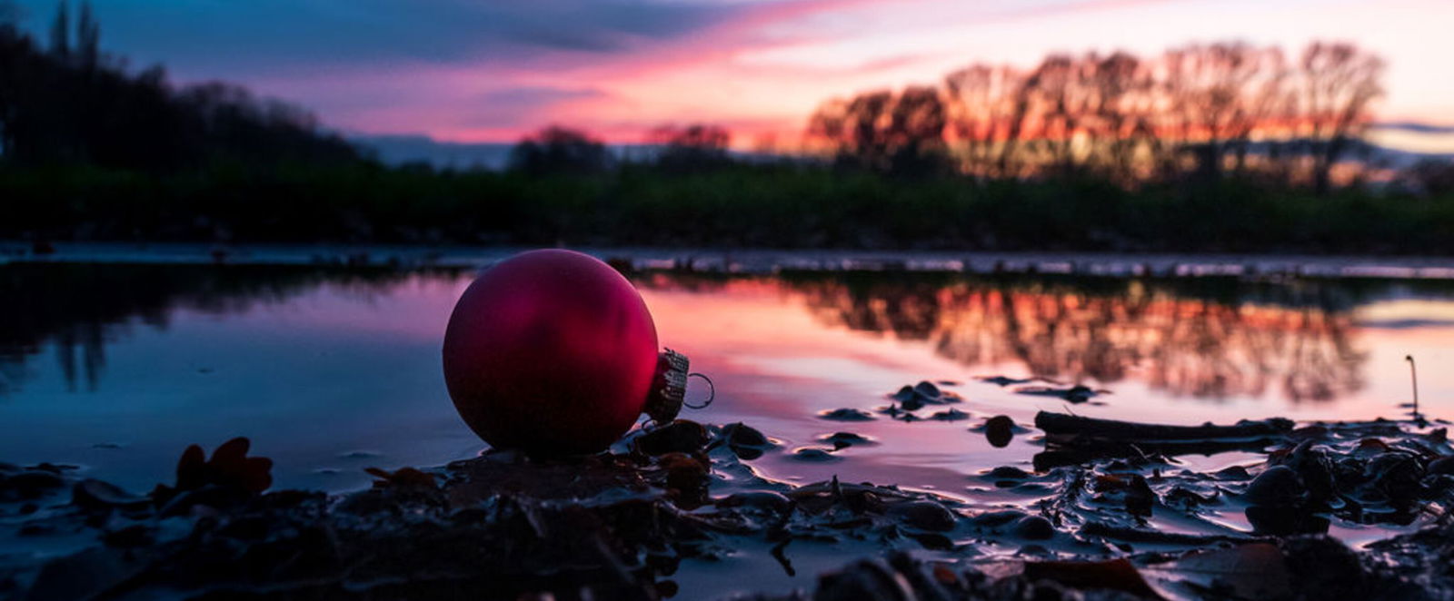 Eine rote Christbaumkugel liegt im Matsch. Im Hintergrund geht die Sonne unter.
