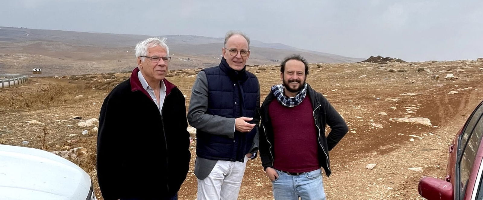Drei männlich gelesene Personen stehen vor einer Geröllbrache im Westjordanland zwischen zwei Autos.