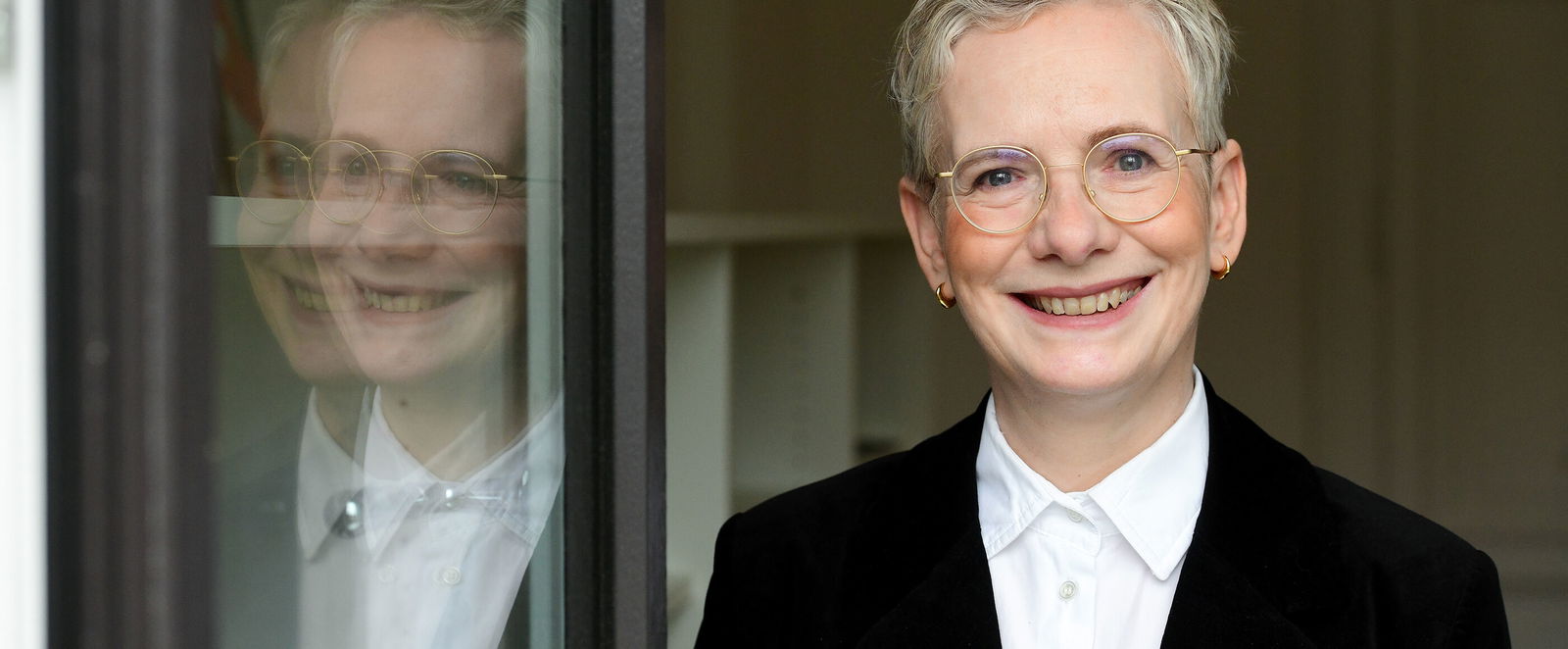 Eine als Frau lesbare Person mit kurzen blonden Haaren blickt in die Kamera. Sie spiegelt sich links in einer Glastür.