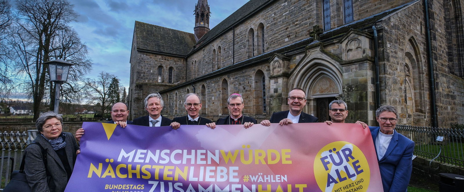 Eine weiblich zu lesende Person und sieben männlich zu lesende Personen halten ein Banner. Im Hintergrund ist eine Kirche zu sehen.