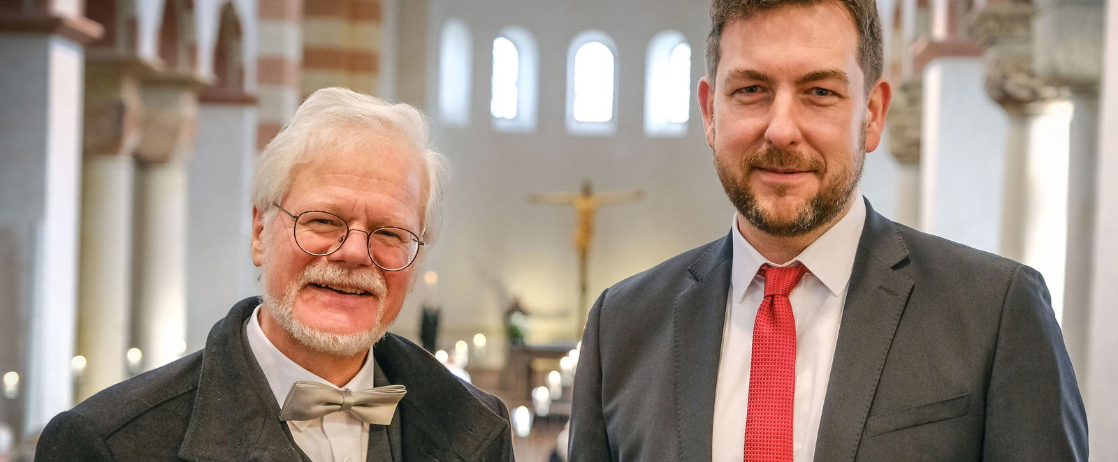 Zwei männlich gelesene Personen, eine älter mit weißem Haar und Brille und Fliege, neben einer jüngeren mit rotem Schlips in einer Kirche.