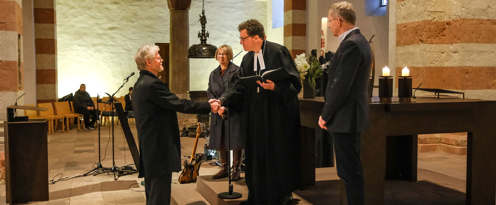 Drei Menschen stehen erhöht vor einer anderen Person. Die mittlere trägt Talar und reicht der entgegenstehenden Person die Hand.