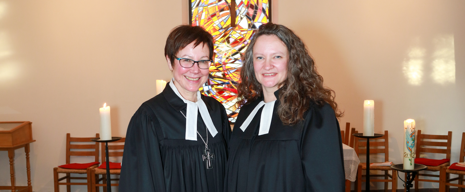 Zwei weiblich gelesene Personen in Talaren stehen vor einem Buntglasfenster mit Kreuz.